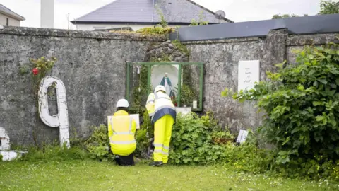 Two workers in yellow high visibility clothing at a memorial left at the site of the  former Mother and Baby Institution in Tuam, County Galway.