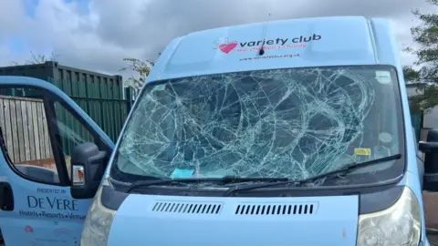 A white van has a badly smashed windscreen wiper. The passenger door is open. Writing reading "variety club the children's charity" is printed above the windscreen.