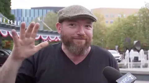 A man with a beard and a grey flat cap gestures with his hands.