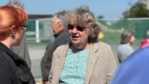 Angela talks to other local residents in front of the site of the proposed warehouse. She is wearing a green top, tweed jacket and sunglasses. The woman she is talking to has red hair, glasses and a black denim jacket.