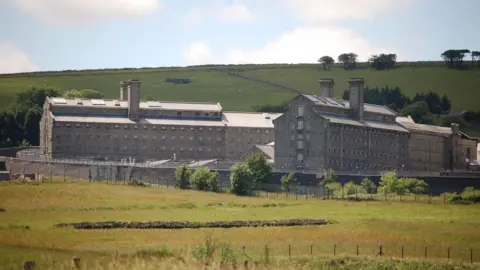 BBC A picture taken from the outside of Dartmoor prison. There is a large grass field to the front of the image. The building is all brick with a number of windows.
