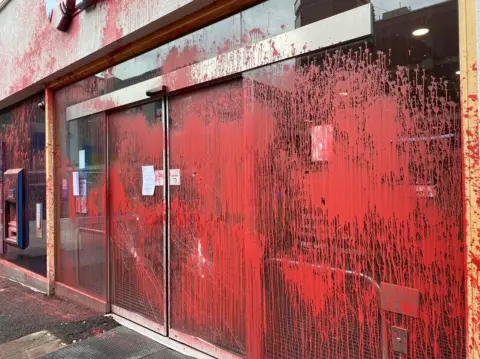 Red paint covering the front of a Barclays bank. The glass doors have been smashed.