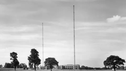A black and white photo shows two large masts above a two storey building, surrounded by trees.