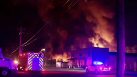 A fire burning in the background with a giant plume of smoke above. Emergency vehicles surround the scene.