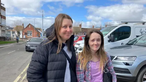 Vikki Irwin/BBC Hannah Goode and Sarah Fox are standing together by a car park. There are buildings and a road to the left of the picture and cars to the right. Hannah is wearing a black puffer coat with a hood and a grey jumper underneath. Sarah is wearing a pink top and pink and green checked shirt. Both are smiling at the camera. 