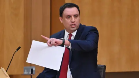 Getty Images Anas Sarwar, who has short black hair, speaks at a podium with a piece of paper in his left hand. He is wearing a dark suit, white shirt and red tie. 