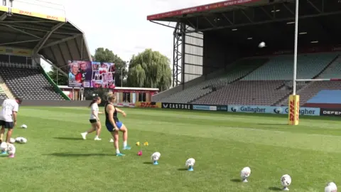 Ella Cromack, wearing a dark T-shirt and blue shorts and light blue boots, kicking a rugby ball. Other rugby balls are lined up ready for her to kick.