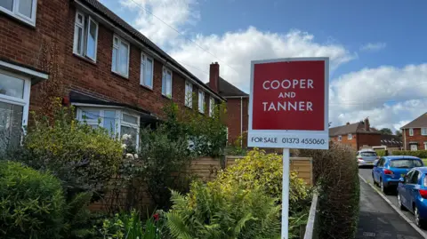 A for sale sign outside a property in Frome. It is a red brick house with several small shrubs in the garden