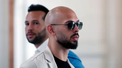 EPA-EFE/REX/Shutterstock Andrew Tate facing to the right, wearing aviator style sunglasses, his head is shaved and he has a dark moustache and beard and wears a black T-shirt and white jacket. His brother is standing next to him facing the other way.