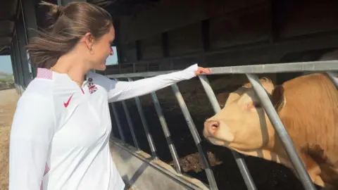 Gemma Tutton, wearing a white GB athletics top, stands in front of a brown cow in a cow shed.