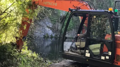 A picture of an orange digger beside a quarry and some trees