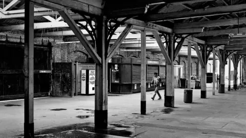 David Pearce A black and white image showing Lowestoft concourse with a man walking through it. It shows beams holding up a roof and rain can be seen dripping down on the top left with a large puddle in the bottom left