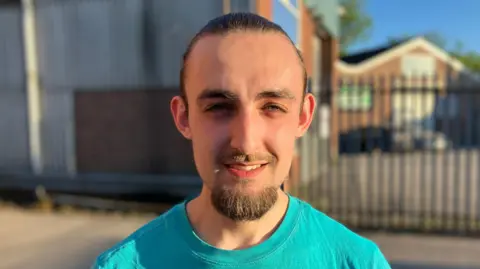 Josh Shepard, a man wearing a turquoise t-shirt, is stood outdoors in the car park of an industrial unit.