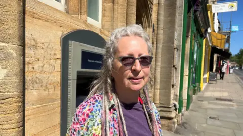 A head-and-shoulders shot of Meig Lambert-Shiels. She is looking directly at the camera and is wearing colourful clothes. She has sunglasses on and there are some old stone buildings in the background.