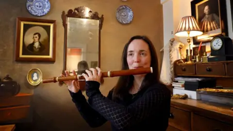 Claire Mann, who has long brown hair is wearing a dark long sleeved top and holding a boxwood flute to her mouth and is standing in a room with traditional furniture and a painting of Robert Burns in the background