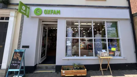 Wychavon District Council The exterior of a white Oxfam charity shop. A small billboard inviting people in stands on the pavement outside, as well as a table with a box of items on it and a box of plants.
