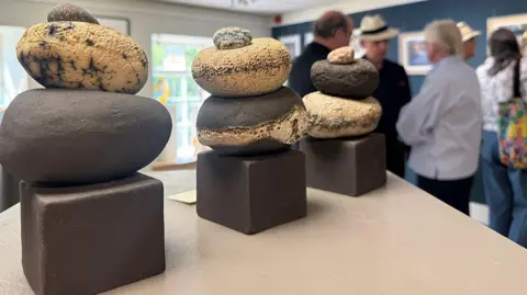 CAROLA RUSH Three clay round sculptures placed on a beige table. There are people in the background talking and looking at other artworks on a wall.