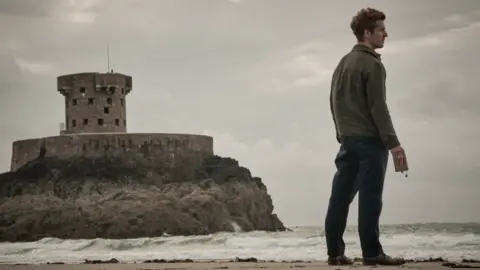 UKTV Damien Molony is holding an empty alcohol flask looking out onto the ocean. He's standing on St Ouen's Bay with La Rocco tower and a moody, grey sky behind him.
