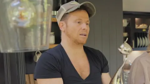 Joe Swash stands in a kitchen looking at a trophy, while wearing a grey cap and black T-shirt
