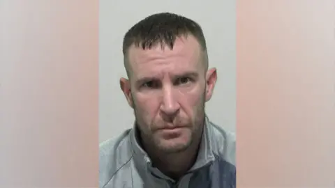 A mugshot of Kevin Belton. He has short, dark brown hair cropped at the sides and a stubbly but relatively neat beard and moustache. He is wearing a grey zipped jacket.