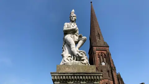 A statue of Robert Burns with a seagull on its head in front of the tower of a sandstone church