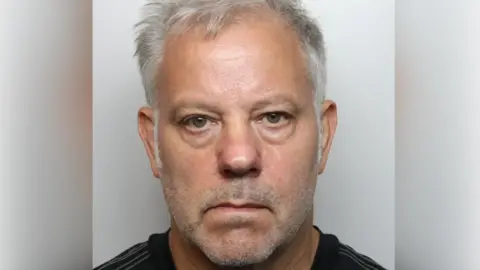 A mugshot of a man with short grey hair and wearing a black top.