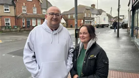 Phil Harrison/BBC A man in a grey hoodie and a woman in a green top and black jacket, stood next to each other on a pavement. The road and some houses can be seen behind them.