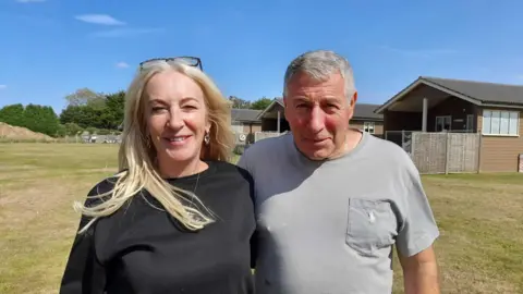 Sharon De Luca and Angelo Dama stand in front of some of their holiday lodges. Ms De Luca, who is smiling, has blonde hair and has glasses on top of her head. She is wearing a black top. Mr Dama has grey hair and is wearing a grey t-shirt. He has his arm around Ms De Luca.  