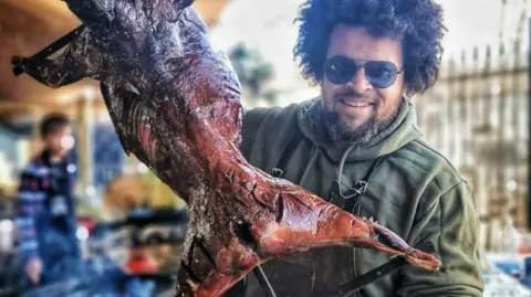Telford and Wrekin Council A man with black curly hair and sunglasses in a green jacket holding up a large piece of roasted meat
