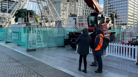 Inspectors pictured on the site of the City Star Flyer on Thursday. One man is in a hi-vis jacket. He is with another man and a woman. In the background, the ride can be seen cordoned off by a green fence.