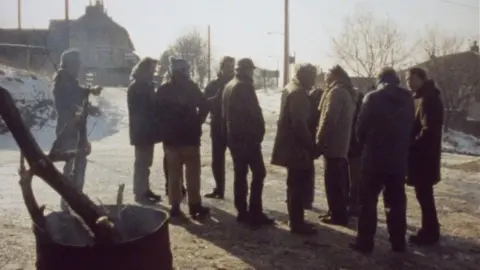 Kjell-Åke Andersson A group of men standing on the road outside the entrance to Oakdale pit. It's winter and there's some snow on the ground. The men are wearing large coats and some are wearing hats and gloves. In the foreground there's an empty oil drum with smoking tree branches sticking out of the top. The background there are some bare trees and the start of a row of terraced houses.