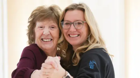 Brenda Grebol smiling at the camera with her dance teacher.
Brenda has mousey brown hair and wearing a dark purple top. Her teacher is blonde and wearing glasses. She is wearing a black blouse