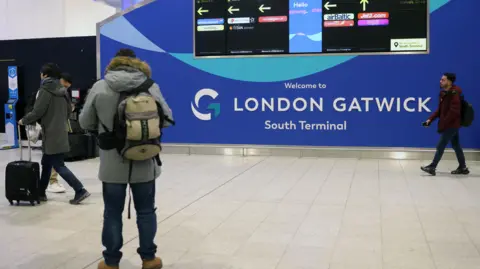 A man with a backpack on stands in front of a wall at Gatwick airport. The sign reads 'Welcome to London Gatwick South Terminal'. A screen shows directions to different airlines.  