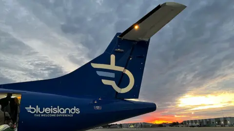 The tail of a Blue Islands aircraft during sunset, with passengers boarding near the rear door. The tail is painted blue and features the airline’s name and slogan, “THE WELCOME DIFFERENCE.”