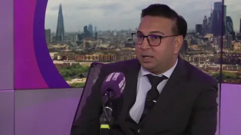 BBC A man sitting at a microphone in the BBC London studio. He is wearing a suit and glasses, with a screen displaying the London skyline behind him.