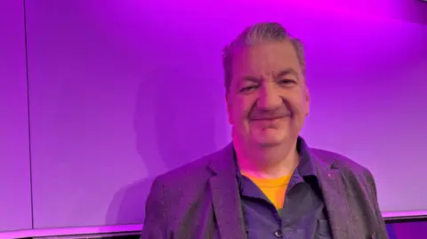 Bill Corcoran has grey hair and is wearing a grey blazer. He is standing in a purple-lit BBC radio studio and smiling.