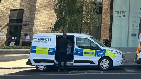 A Metropolitan Police "Forensic Services - Crime Scene Investigation" van parked on the street outside a modern brick building labeled "Tiller & Lock." An officer seen from behind is reaching into the open side door of the van.