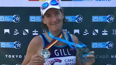 Gill Punt Gill has a short brown bob and is wearing a white cap with blue sunglasses on top of it. She is wearing a blue, purple and white vest that says "Gill, cancer research". She has two medals around her neck and is holding them up. On the last is a ribbon with the German flag on it, and the medal is silver with a person cheering on it. On the right is the marathon majors medal, which is a large circular silver medal made up of six smaller circular medals with a blur ribbon