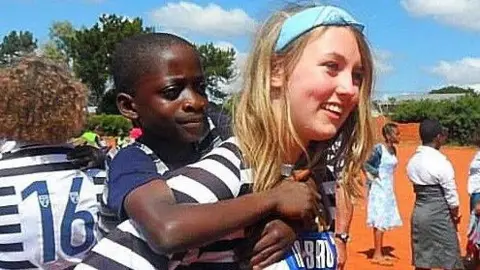 Allan family Katie in Malawi gives a piggy-back to a little boy during a football match. She is smiling broadly, wearing a football strip - other youngsters behind her are also wearing the strips and some girls stand in the background in the school playground.