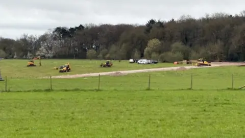 A green field with a barbed wire fence running through the middle. At the back of the field, there are several diggers roam around and piles of gravel are stacked up.