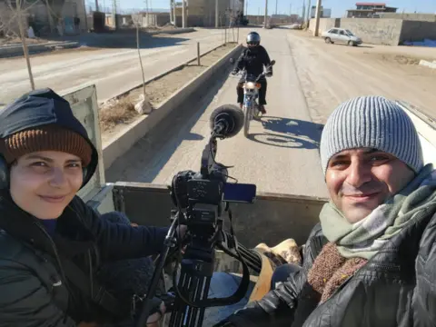 Gandom Films A woman and a man sit in a pick-up truck filming a motorcyclist riding along the road out the back, a camera in between the two people who are both wearing hats and coats.