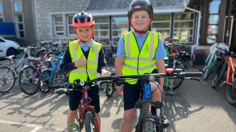 Two children wearing helmets and high-visibility vests, standing with their bicycles in front of a bike rack that holds several other bicycles. In the background, there's a stone building with lots of glasses windows.