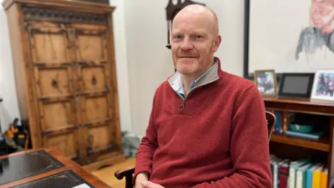 BBC Deputy Gavin St Pier looking towards the camera wearing a red jumper with a blue collared shirt. 