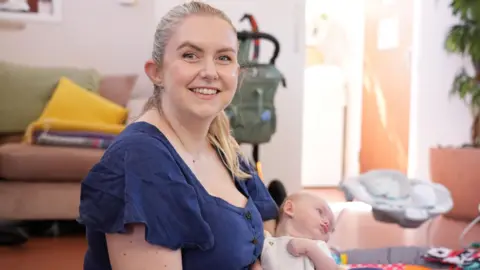 Becks Wilson with her baby Dylan in her arms. Her blonde hair is tied up and she is looking at the camera, smiling. They are sat on the floor in a well lit room. A baby bouncer chair can be seen in the background to the right of the image. To the left there is a sofa with blankets piled on top of it.