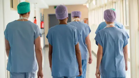 Five medical staff members walk through a hospital corridor. Their backs are to the camera. They are wearing blue medical scrubs and paper hats.