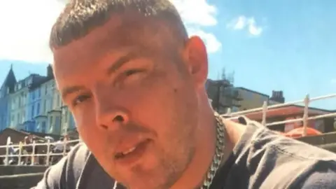 West Yorkshire Police A man with short brown hair and facial stubble, He is wearing a grey t-shirt with a heavy silver necklace. 