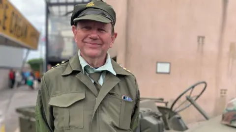 Phil Cunliffe/BBC A man wearing a dark green army uniform and hat stands smiling in front of a camouflage coloured vehicle.