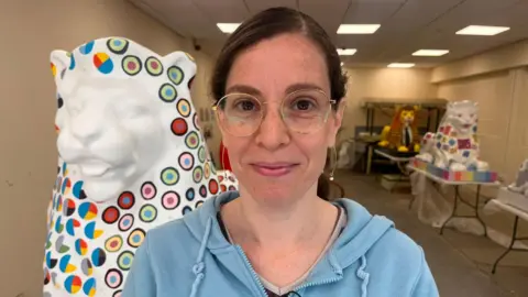 Or is smiling at the camera. She has brown hair that is pulled back into a ponytail. She is wearing a blue zip-up hoodie. She has glasses on which have a gold frame. Behind her are a number of painted lions. She is stood next to hers which she designed. It is a white in colour with a number of data graphs painted on it.