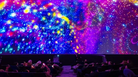 A large group of people recline in seats inside a dark planetarium-style dome while a vivid, colourful projection fills the entire curved ceiling. The display shows swirling patterns of bright neon colours - pinks, blues, yellows, and purples - resembling galaxies, stars, and abstract light bursts. Several audience members hold up phones to capture the immersive light show.