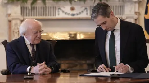 PA Michael D Higgins - an older man with white hair wears rimless glasses is sat at a desk with Simon Harris. He has short dark hair and is signing a piece of paper. Bother men are wearing dark suit jackets, white shirts and ties.